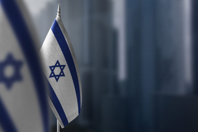 Photo: Small flags of Israel are displayed in front of a blurry background of a city.