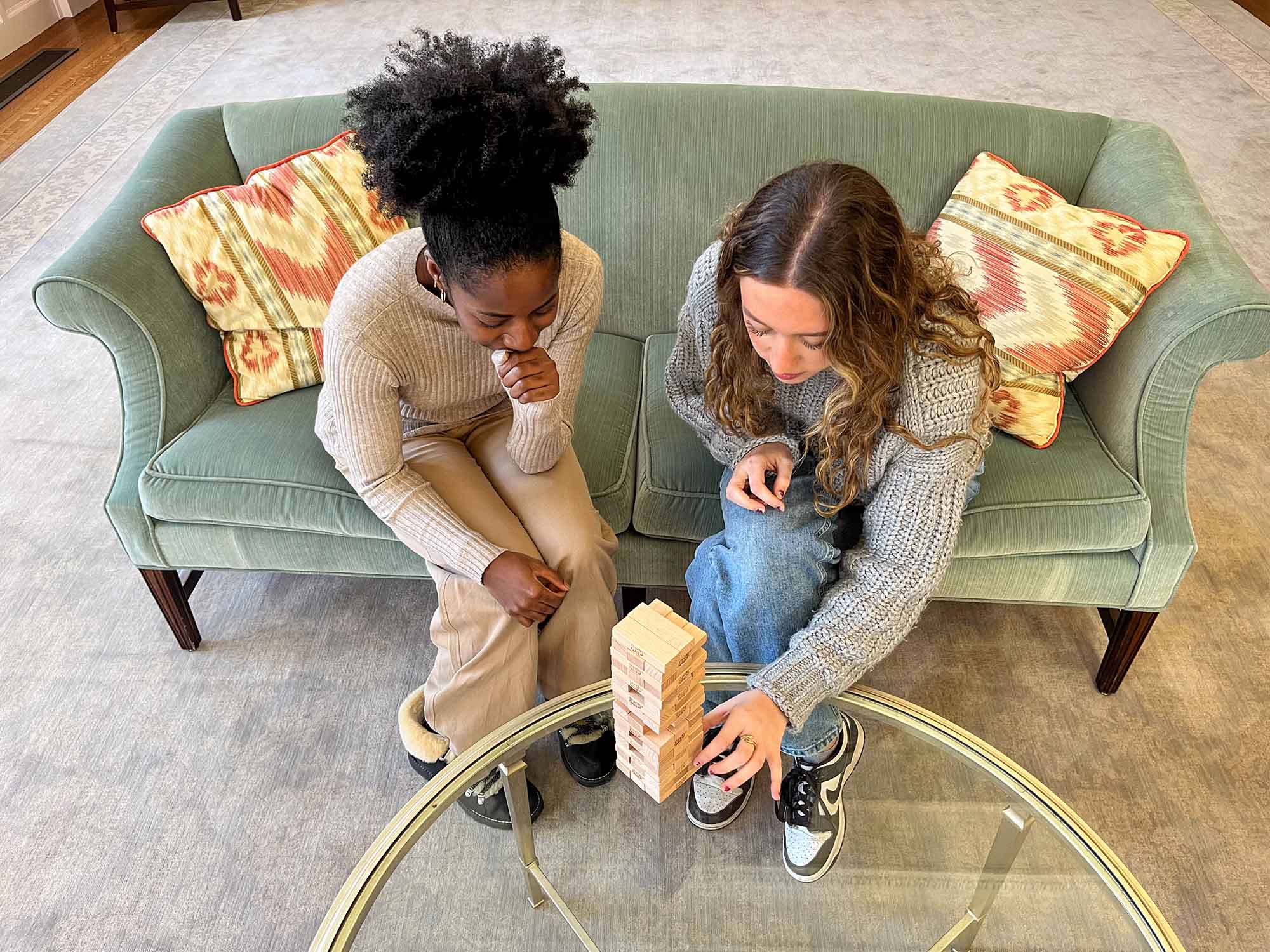 Photo: A Black woman with hair tied up in an afro puff and wearing a an oatmeal-covered sweater and tan pants sites to the left of a young white woman with long brown hair and wearing a grey sweater and jeans. They both sit on a muted green couch and play a game of Jenga.