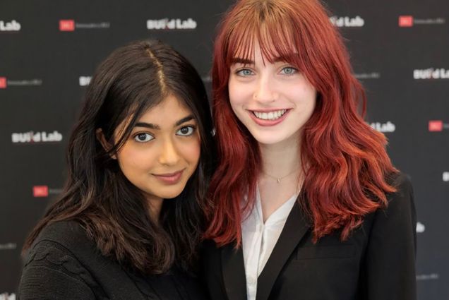 Photo: Prianna Sharan (left) and Remi Chester pose in front of a black BU Build Lab backdrop. (left) Prianna, a South Asian young woman with long black hair and wearing a black top poses closely next to Remi, a young white woman with deep auburn red hair and wearing a white collared shirt and black blazer.