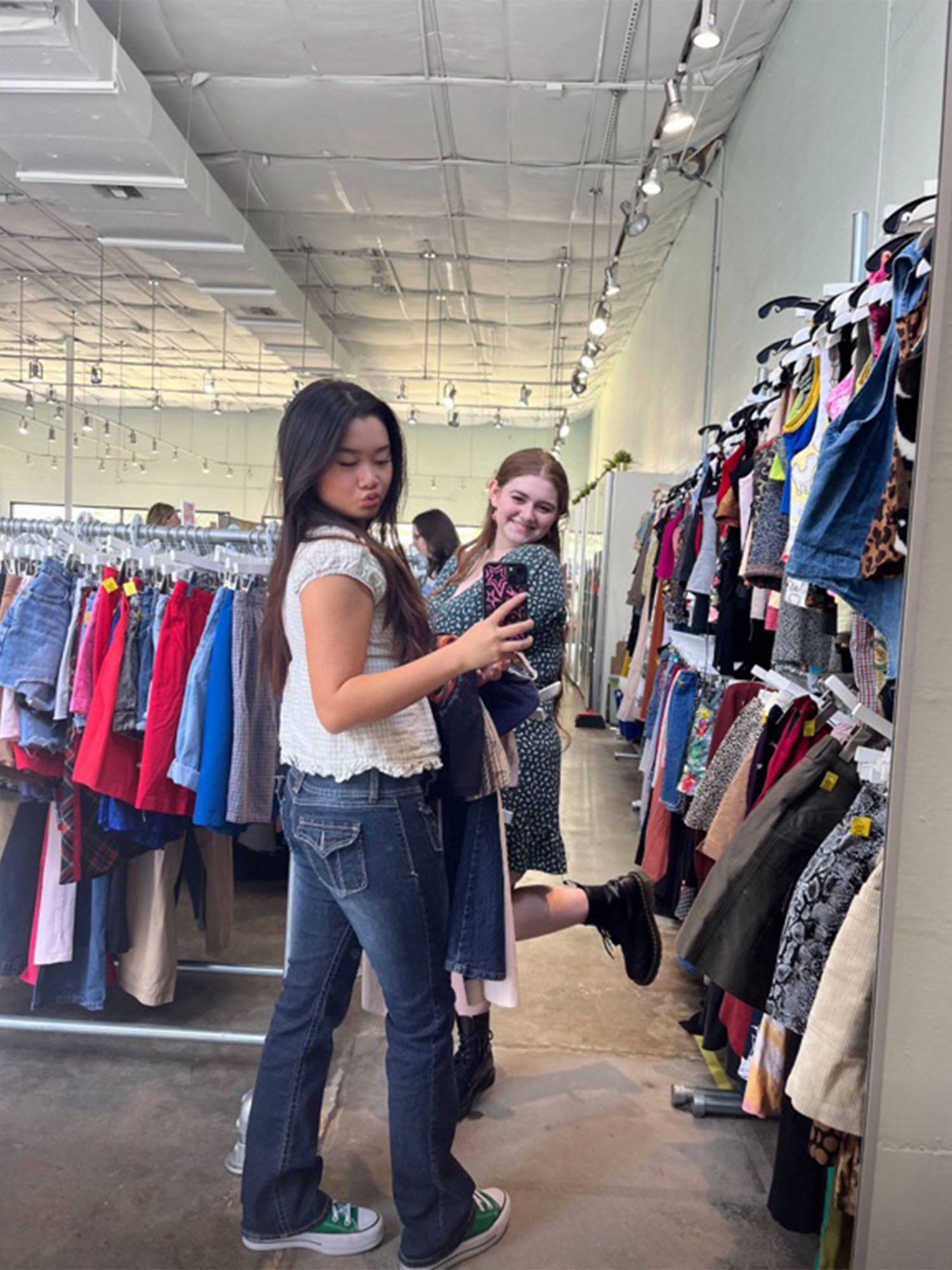 Photo: Two college students shopping for women's clothing at a thrift store. They are standing in front of a mirror taking a selfie, with clothing racks surrounding them