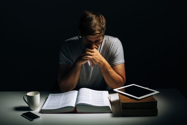 Photo: A stock image of a man studying with books, papers, and additional educational accessories spread out on a table. he is shadowed by a spotlight to his right, as he contemplatively looks over everything on the spread.