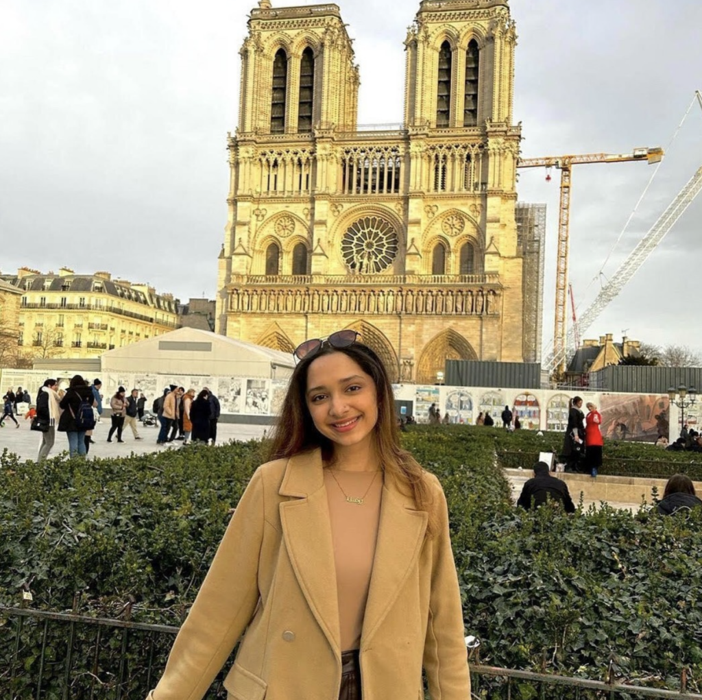 Boston University student Rukshana Khan. She is wearing a tan shirt with a tan jacket over it, smiling at the camera. She is abroad in this photo, standing in front of an intricately designed gothic style building. 