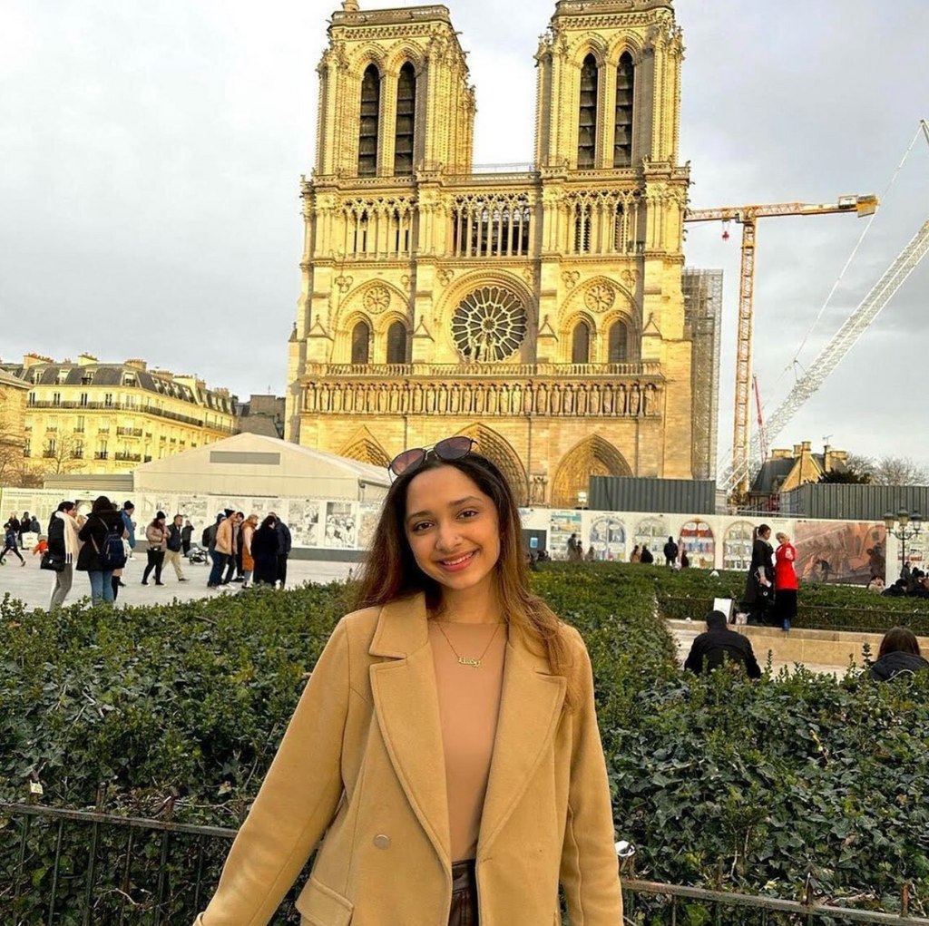 Boston University student Rukshana Khan. She is wearing a tan shirt with a tan jacket over it, smiling at the camera. She is abroad in this photo, standing in front of an intricately designed gothic style building.