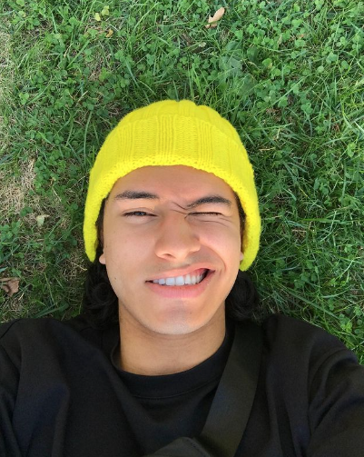 This photo is courtesy of Boston University student, Andrés Marquez Santacruz. He is laying on green grass, wearing a yellow hat and black shirt, squinting at the camera.