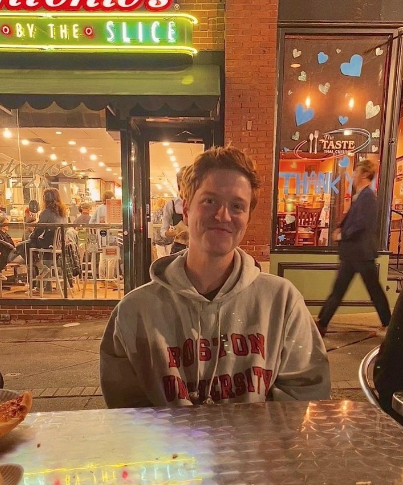 Photo is courtesy of Joey Russoniello. He is outside at a restaurant at dusk wearing a grey Boston University hoodie. He is smiling with his mouth closed and has ginger hair. 