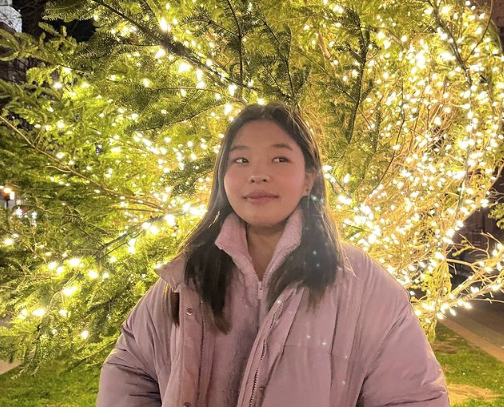 This photo is of BU student Esther Yang. She is standing in front of a fairy light wrapped evergreen tree. She is wearing a pink jacket and has her hands in her pockets. She's looking off into the distance with a small smile on her face. 