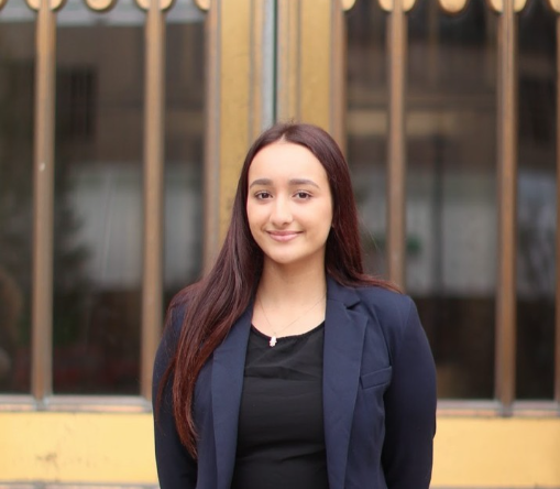 This photo is courtesy of Boston University student Sarah Benhalima. She is wearing a professional blazer and is smiling. Her hair is down and straight and she is standing in front of a building. This is a professional headshot.
