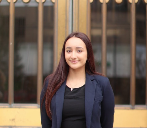 This photo is courtesy of Boston University student  Sarah Benhalima. She is wearing a professional blazer and is smiling. Her hair is down and straight and she is standing in front of a building. This is a professional headshot. 