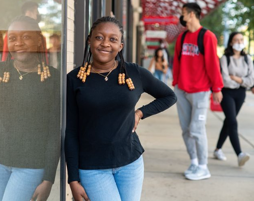 This photo is of Boston University student, Brianna Bourne. She is wearing a black longsleeve with blue jeans and she is leaning against a building. There are students blurred and walking in the background. She is smiling for the photo. 