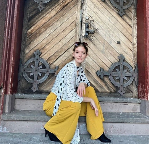 This photo is of Boston University student Anna Rafferty. She is wearing yellow pants and a patterned black and white top. Shes sitting on steps and is leaning her weight on one knee. 