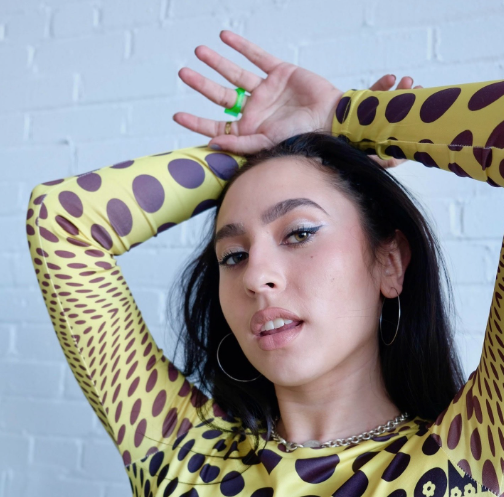 This photo is of Boston University alum, Isadora "Izzy" Bernasconi. She is wearing a yellow and black patterned shirt with her hands above her head. She is looking at the camera with her mouth slightly parted.