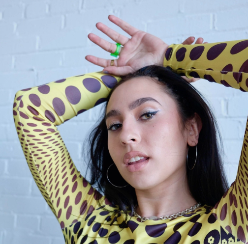 This photo is of Boston University alum, Isadora "Izzy" Bernasconi. She is wearing a yellow and black patterned shirt with her hands above her head. She is looking at the camera with her mouth slightly parted. 