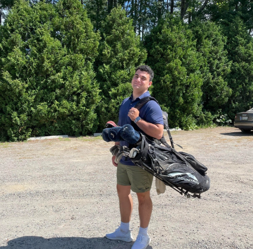 This photo is of Boston University student, Andrew Ciali. He is standing inn front of green trees, and is in golf attire. He's holding a golf bag on one shoulder, standing to the side, smiling at the camera. 