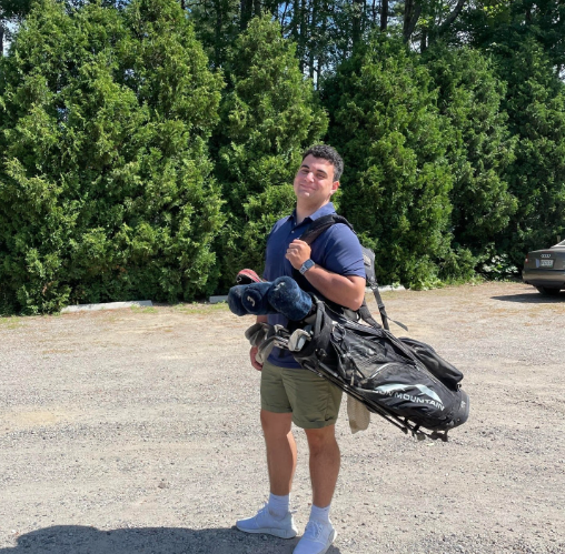 This photo is of Boston University student, Andrew Ciali. He is standing inn front of green trees, and is in golf attire. He's holding a golf bag on one shoulder, standing to the side, smiling at the camera.