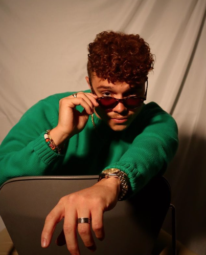 Boston University student Brendon Jones. He is wearing a green sweater with gold jewelery, leaning forward in a chair. He has sunglasses on and he is lowering them to look at the camera with one hand. His other arm is sticking forwards.