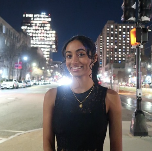 Boston University student Navya Kotturu. She is in the city with buildings lit up behind her at dusk. She is wearing a black tank top and has her hair up. She is smiling at the camera. 