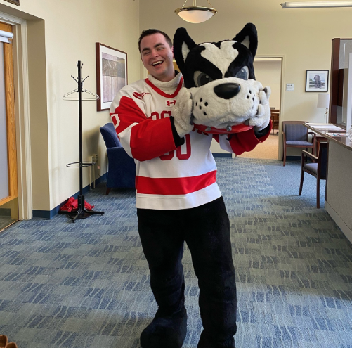 Boston University student Matthew Pruitt. He is wearing the Boston University mascot Terrier costume, holding the head to the side, smiling at the camera. He is in an office building with blue carpeting.