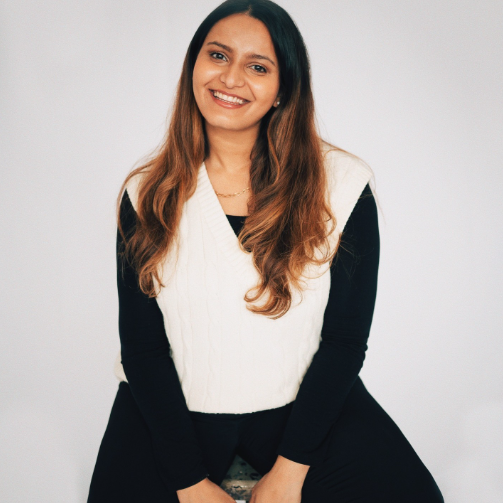 Boston University student Paulomi Mukherjee. She is sitting on a stool wearing all black with a white sweater over it. She is smiling and has her hair down, looking towards the camera. She is in front of a white background. 