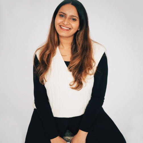 Boston University student Paulomi Mukherjee. She is sitting on a stool wearing all black with a white sweater over it. She is smiling and has her hair down, looking towards the camera. She is in front of a white background.