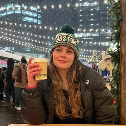 Boston University student Noa Blitz . She is wearing a beanie hat on top of her head with a black puffer coat on. She is sitting at a restaurant outdoors holding up a hot chocolate. She is looking off into the distance with a soft smile on her face. There are yellow fairy lights above her.
