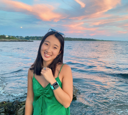 Boston University student Sofia Lee. She is standing on a beach while the clouds have a pink hue to them. She is wearing a green satin dress and is smiling at the camera. 