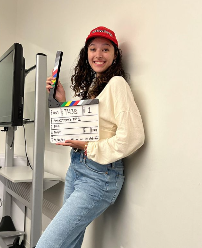 Boston University student Maggie Borgen. She is wearing blue jeans, a cream longsleeve top, and a red baseball cap. She is leaning against a white wall while holding a directors take in her hands. 