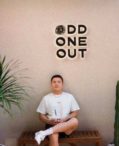 Boston University student Bill Yao. He is sitting cross legged on a bench in front of what appears to be a cafe. The wall it tan and there is a palm tree to his left. He is in shorts and a white t shirt.