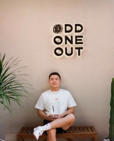 Boston University student Bill Yao. He is sitting cross legged on a bench in front of what appears to be a cafe. The wall it tan and there is a palm tree to his left. He is in shorts and a white t shirt. 