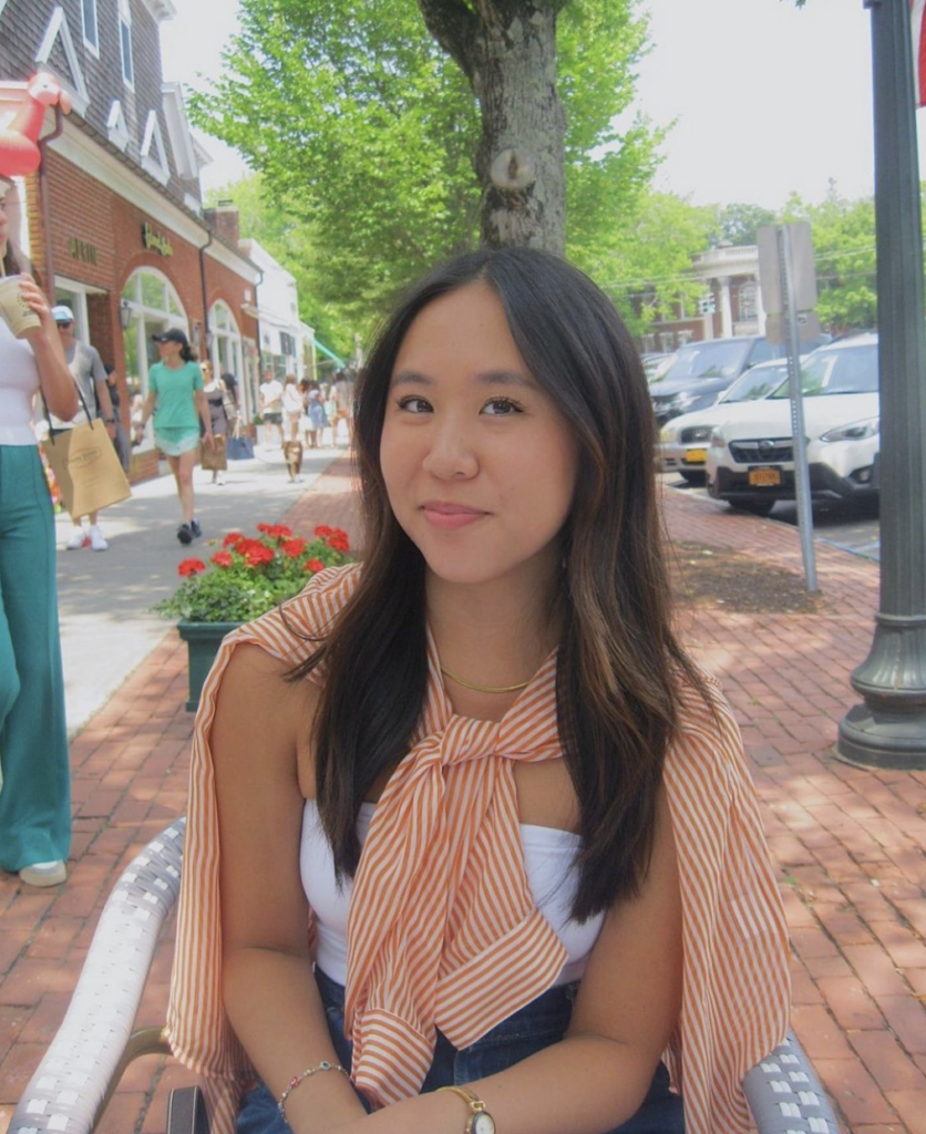 Boston University student Miri Chan. She is sitting in a chair outside, on a brick road. She is wearing a white tank top with a striped orange shirt tied around her neck. She is smiling at the camera. 