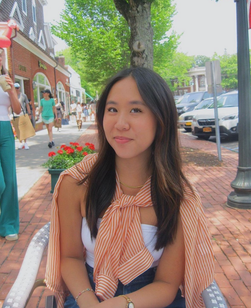 Boston University student Miri Chan. She is sitting in a chair outside, on a brick road. She is wearing a white tank top with a striped orange shirt tied around her neck. She is smiling at the camera.
