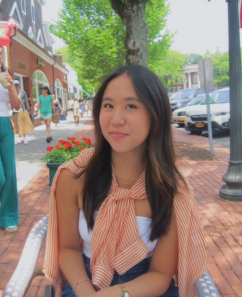 Boston University student Miri Chan. She is sitting in a chair outside, on a brick road. She is wearing a white tank top with a striped orange shirt tied around her neck. She is smiling at the camera. 