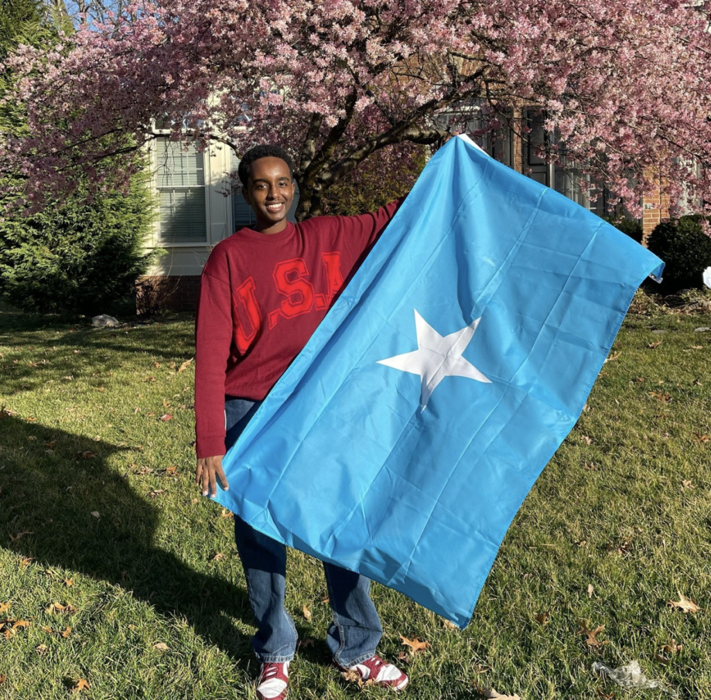 Boston University student Wahaaj Farah. He is outside on green grass, with a cherry blossom tree to his right. He is standing with a Somali flag in his hands. He is wearing a red USA sweater and blue jeans, smiling at the camera.  