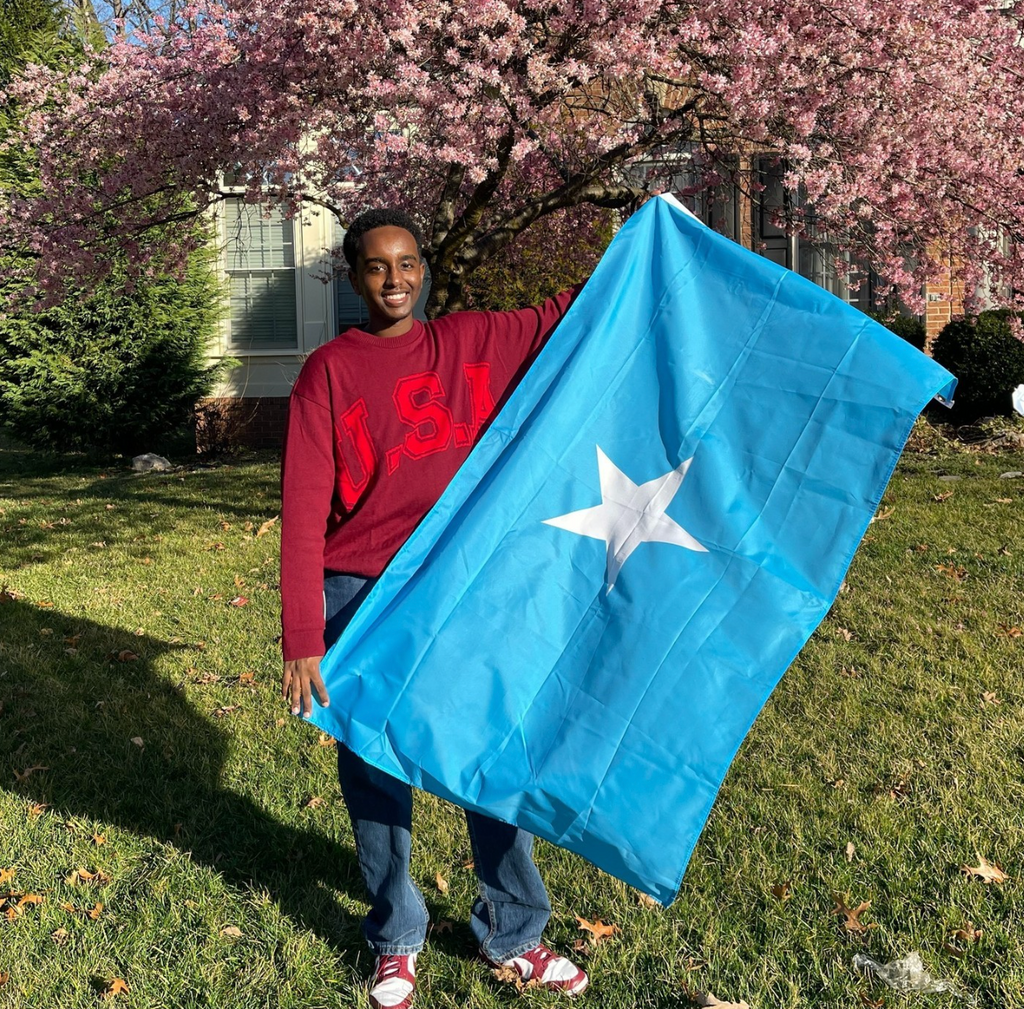 Boston University student Wahaaj Farah. He is outside on green grass, with a cherry blossom tree to his right. He is standing with a Somali flag in his hands. He is wearing a red USA sweater and blue jeans, smiling at the camera.