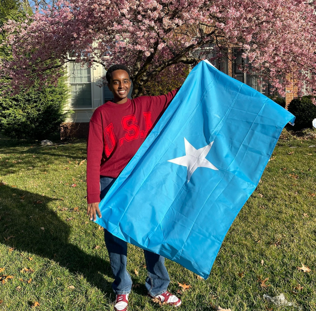 Boston University student Wahaaj Farah. He is outside on green grass, with a cherry blossom tree to his right. He is standing with a Somali flag in his hands. He is wearing a red USA sweater and blue jeans, smiling at the camera.  