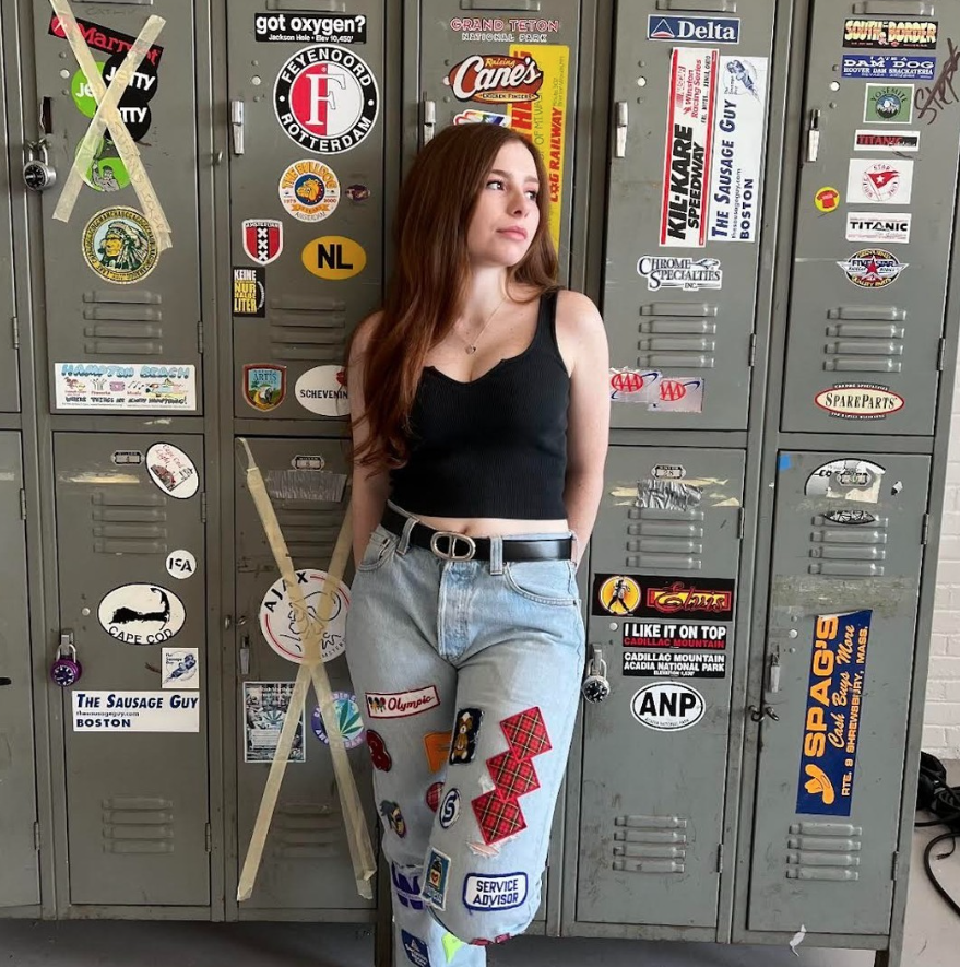 Boston University student Tabitha Fortner. She is wearing patch worked jeans and black tank top. She is leaning against lockers that are covered in stickers. She is looking off into the distance, to the right. 