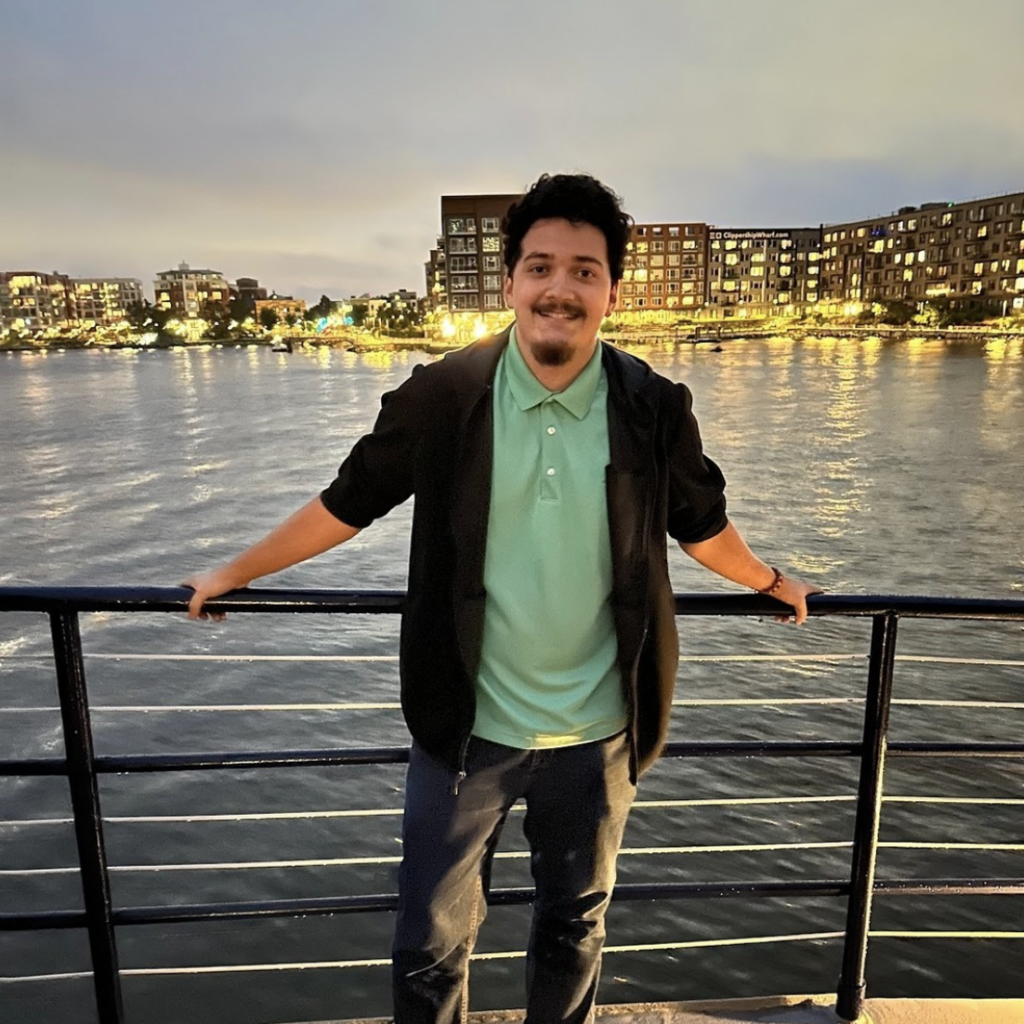 Boston University Student Vincent Semidey. He is wearing a green polo with a black cardigan over it with black jeans. He is standing on a pier while the sun is setting. There is blue water behind him with lights in the distance. He is holding onto the railing behind him, smiling at the camera.