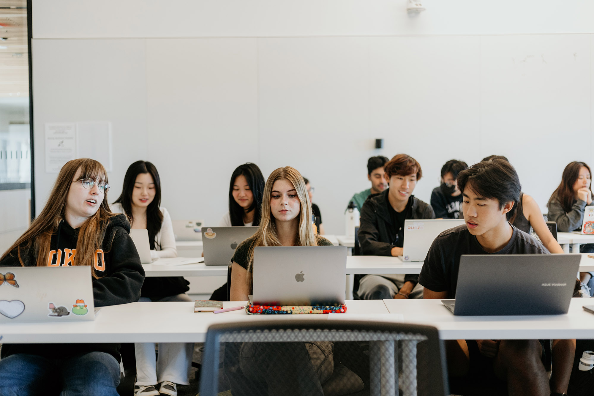 Photo: A classroom of students sitting at long tables arranged in rows, work on their laptops in a bright BU classroom.