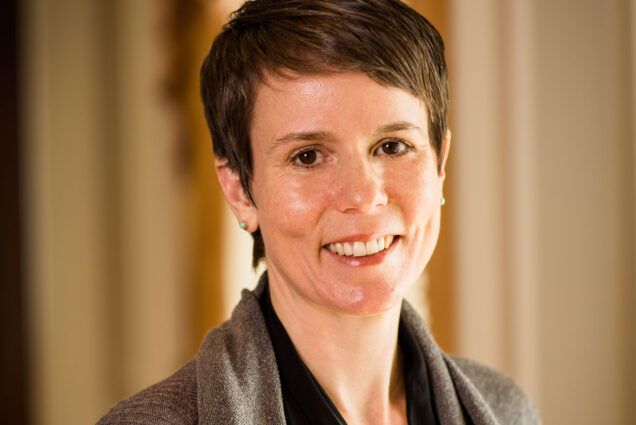 Photo: Headshot of Tracy Schroeder, a white woman with short brown hair and wearing a black blouse and brown cardigan sweater. She smiles in front of blurred office background.