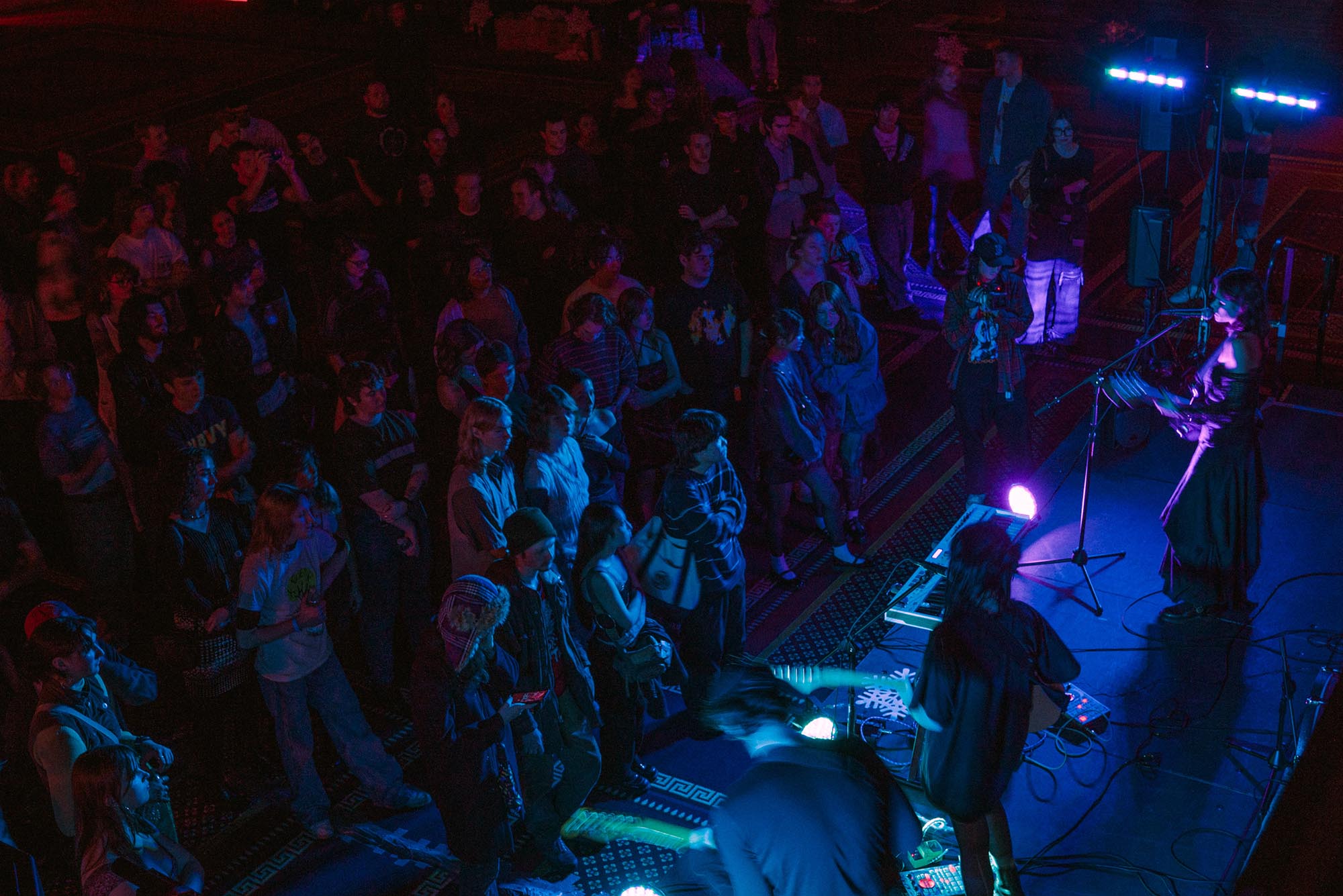 Photo: Overhead shot of a band playing to a large crowd in a medium-sized band space. Three people stand and perform on a stage as the crowd watches in a dark room lit by blue and purple lights.