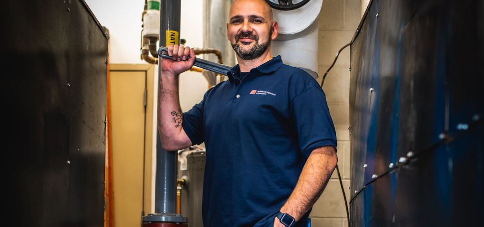 Photo: A man in jeans and a t shirt holding a large pipe wrench on his shoulder, in what appears to be a boiler room