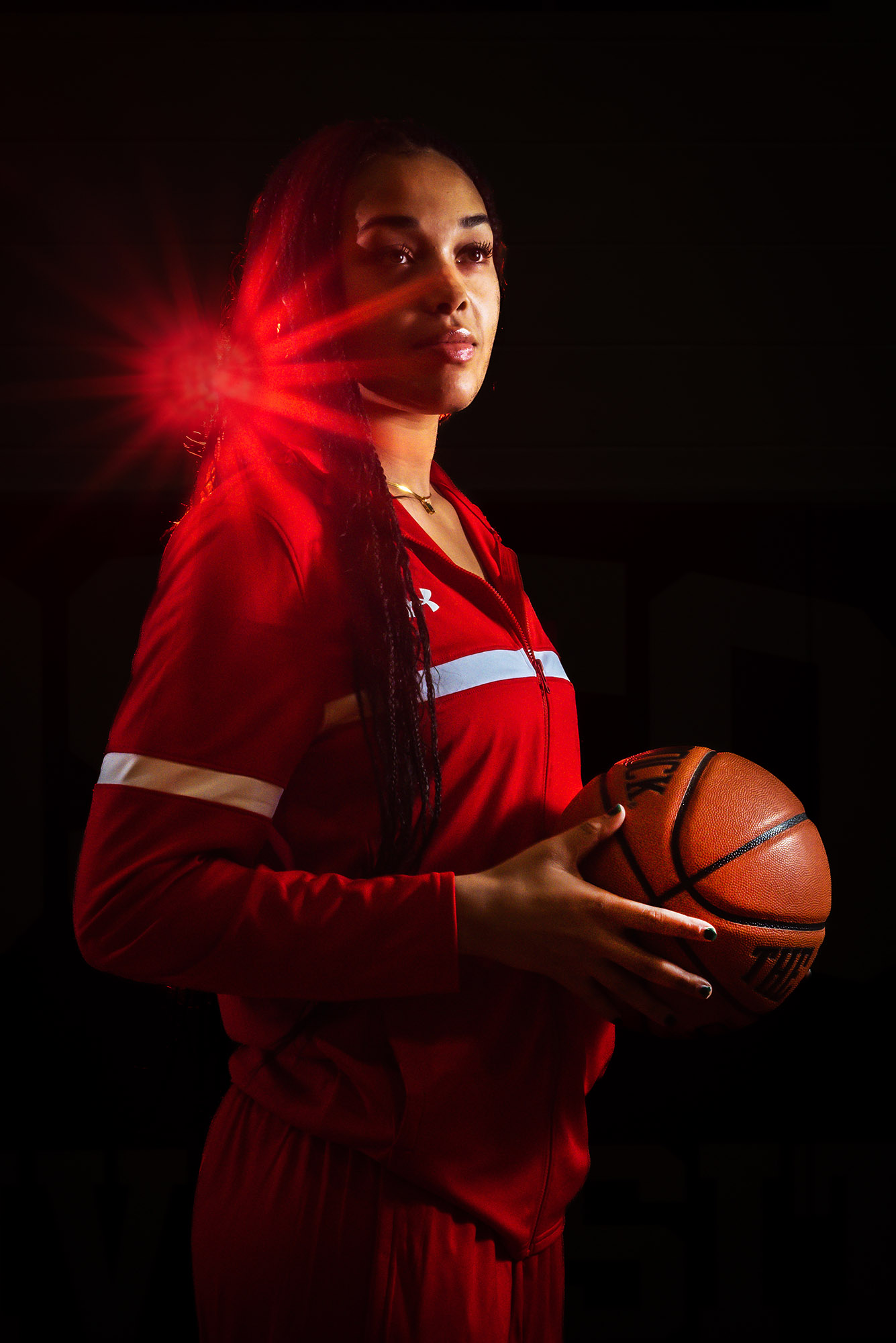 Photo: Women’s basketball player, Caitlin Weimar, poses for a photo with her red uniform. She holds a basketball, and stares to the right. A bright, red light shines behinds her head.