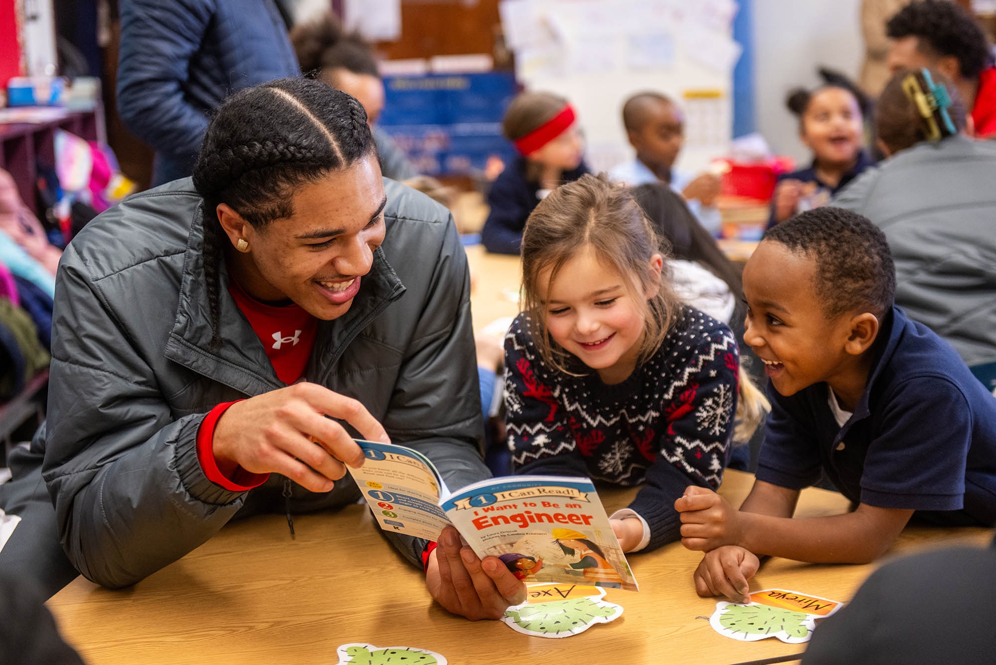 Photo: BU basketball athlete Matai Baptiste (CAS’27), at left, reads a book about engineering to kindergartners. They sit at a small table, the two kids to his right are engaged and radiate joy.