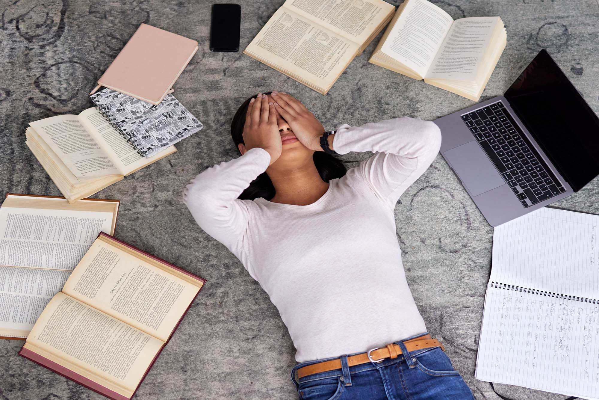 Photo: A person wearing a white top and jeans lies on the floor with hands over face in an exasperated manner. They lie among various open books, notebooks, and laptop.