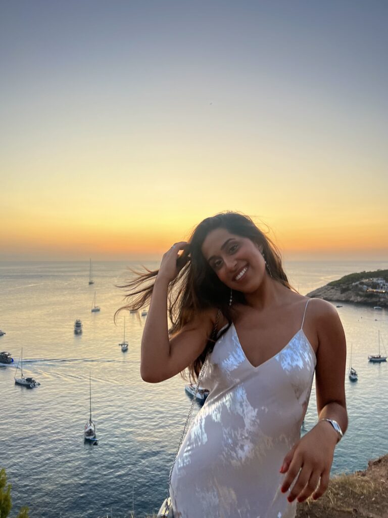 Boston University student Aananya Asnani. She is wearing a white satin dress standing in front of a body of water while the sun is setting behind her. She is somewhere tropical and there are boats in the water.