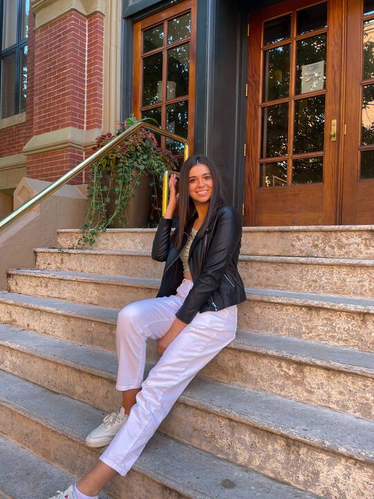 Boston University student Sophia caffrey. She is sitting on white steps in front of wooden doors. She is wearing white pants and a black leather jacket. She has one knee up and her other leg stretched. One of her hands is fixing her hair while the other is resting on her lap. 