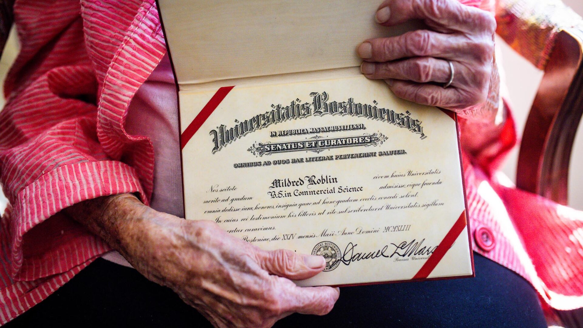 Photo: A pair of wrinkled hands hold an old Boston University Diploma, with red casing and formal letterhead