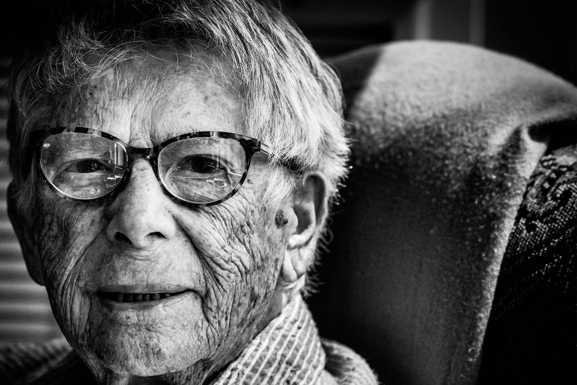 Photo: an up-close portrait of a centenarian wearing glasses and a sweater. Photo is black and white