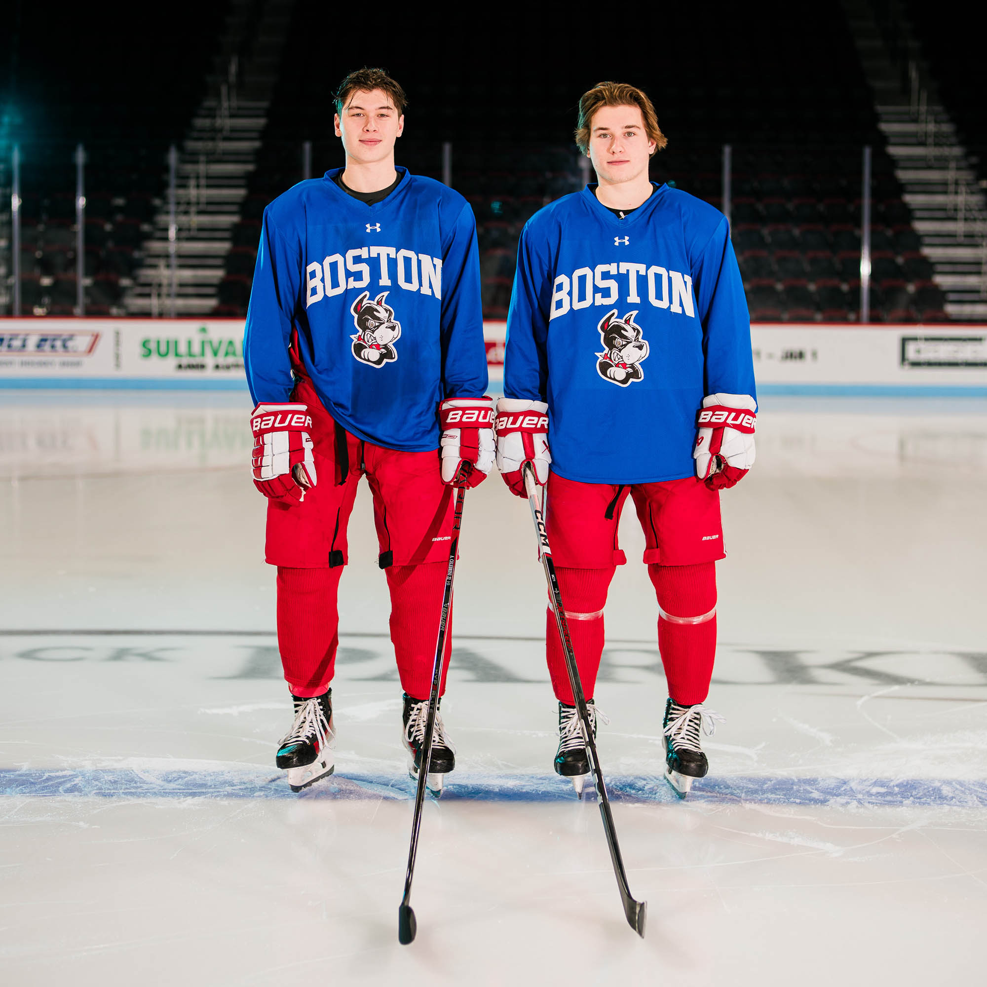 Photo: Two hockey players pose standing on the ice facing the camera. They wear a blue practice jersey and the red issue hockey pants.