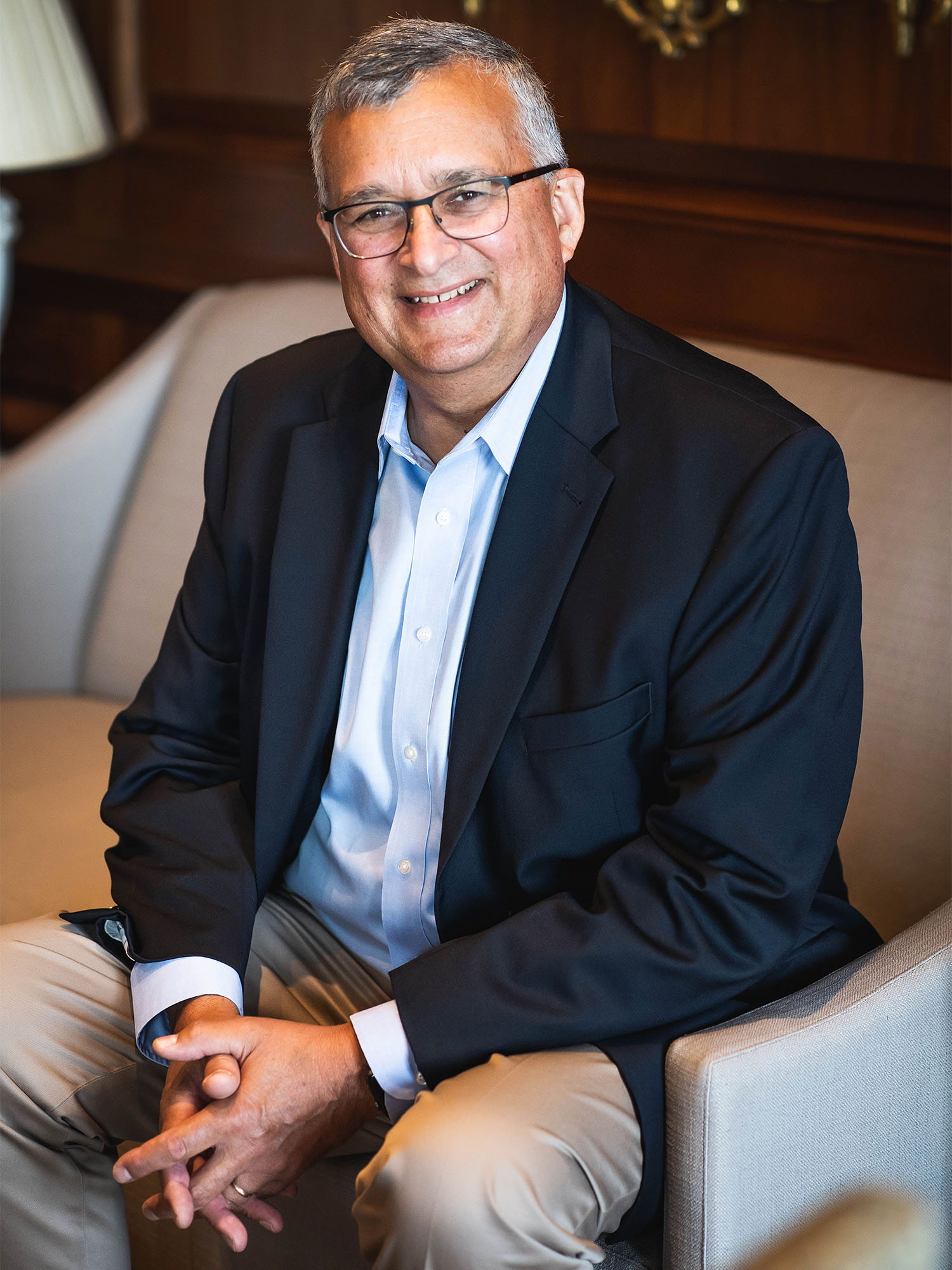 Photo: A middle aged man wearing glasses with a smile on his face sits in a high class couch, wearing a black suit and collared shirt.