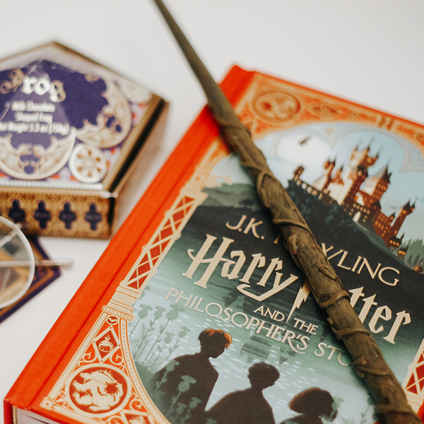 Photo: Overhead shot of the book "Harry Potter and the Philosopher's Stone". Photo shows book on a white table with a brown wand lying on top of it. Glasses and a Bertie Bott's beans container can be seen to the left of the book.